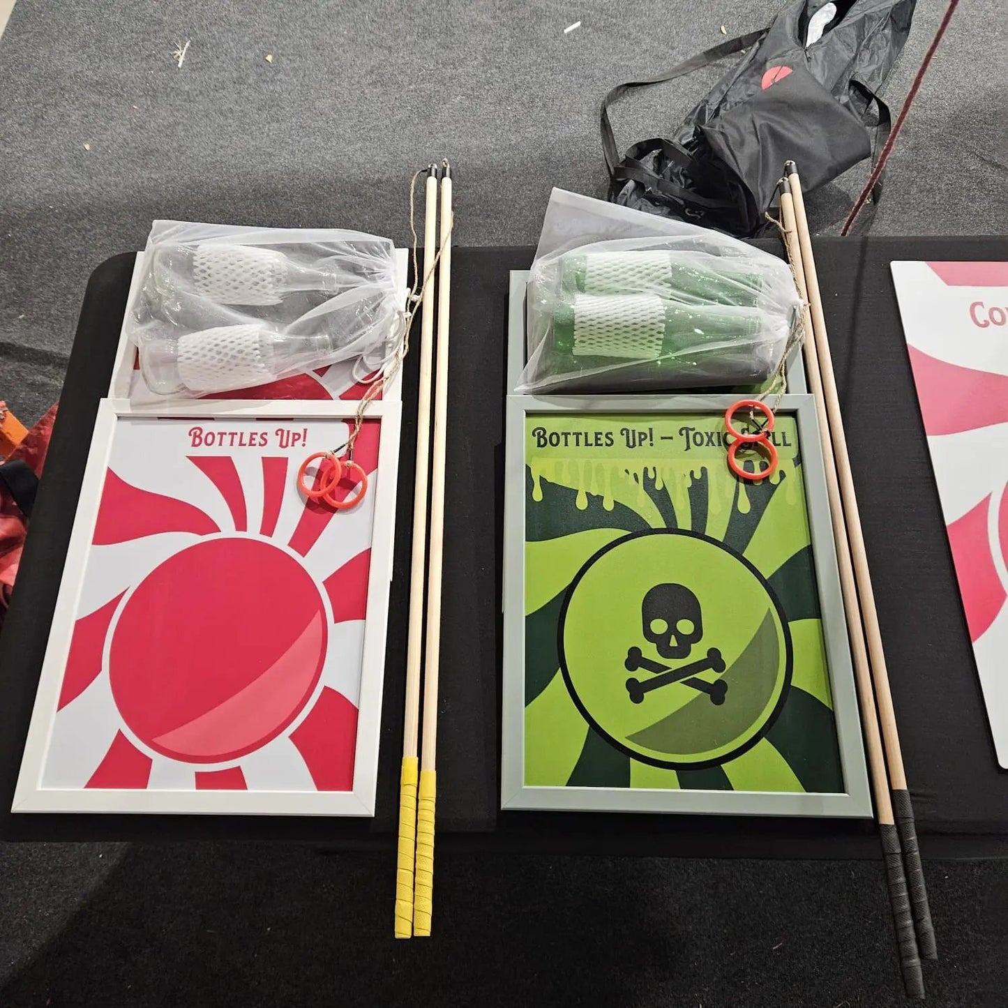 Two game cards with sticks on a black surface, one labelled 'Bottles Up!' with a red circle and the other labelled 'Toxic' with a green skull and crossbones design.